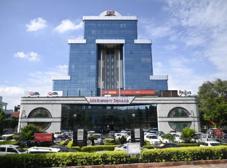 This is a JMD Regent Square building showcasing a straight-on, wide front view of its multi-tier blue glass facade, highlighting prominent tenant signage like HSBC, Kotak, and Regus, alongside a busy front parking lot and manicured green hedges.
