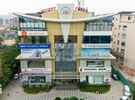 This is a JMD Regent Plaza building showcasing a straight-on, elevated front view of its multi-story commercial facade, highlighting prominent corporate tenant signage, retail showrooms on the ground floor, and a paved entry courtyard.