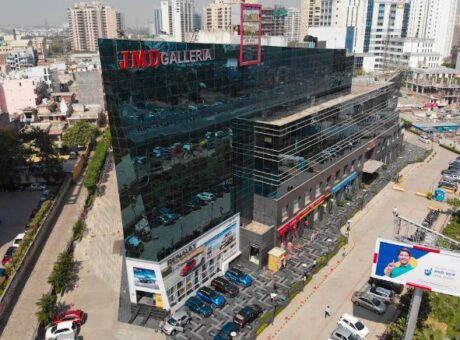 This is a JMD Galleria building showcasing a high-angle drone view of its unique, angular glass-facade structure, highlighting the ground-floor retail showrooms, a multi-car paved parking layout, and the surrounding commercial neighborhood.