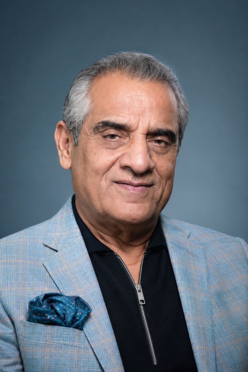 Professional headshot of Mr. Sunil Bedi, Founder of JMD Group, wearing a light blue patterned blazer over a black zippered shirt against a blue background.