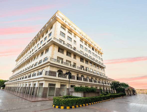 This is a JMD Suburbio 67 building showcasing a low-angle corner view of its multi-story commercial structure against a sunset sky, featuring ground-floor glass retail showrooms, wrapped balconies with black iron railings, and a neatly bordered green hedge perimeter.