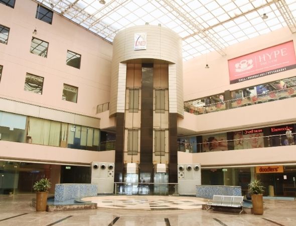 This is a JMD Galleria building in Sector 48 Gurugram showcasing a ground-level view of the central commercial atrium, featuring an indoor cafe with sitting arrangements, a blue mosaic-tiled water fountain, a small decorative holiday tree, and a modern capsule elevator tower in the background.