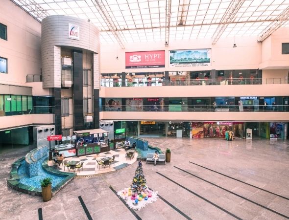 This is a JMD Galleria building in Sector 48 Gurugram showcasing an elevated view of the inner commercial atrium, featuring a central cafe seating area bordered by a blue tiled water feature, a small decorated festive tree, and vibrant brand signage along the upper corridor tiers.