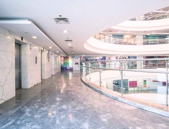 This is a JMD Megapolis building on Sohna Road Gurugram showcasing a wide office passage and elevator lobby zone, featuring clean white modern interiors, recessed ceiling lighting, and a view overlooking the central commercial atrium ring.