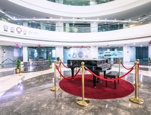 This is a JMD Megapolis building on Sohna Road Gurugram showcasing a luxury ground floor reception lobby entrance, featuring a premium black grand piano placed on a red circular carpet with gold stanchion ropes, surrounded by polished marble flooring and a curved architectural layout.