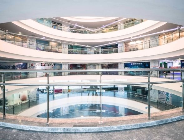 This is a JMD Megapolis building on Sohna Road Gurugram showcasing a wide perspective of its upper-floor interior corridor, featuring a sleek curved glass railing overlook, bright modern ceiling panel lights, and polished flooring reflecting the multi-story commercial atrium.