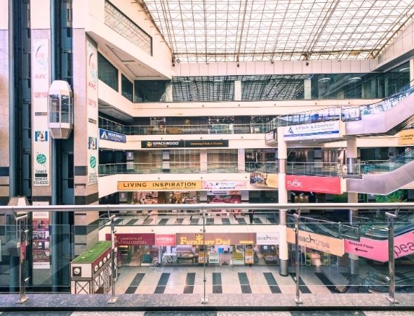 This is an interior architectural shot inside JMD Regent Arcade on MG Road Gurugram, taken from an upper floor railing overlooking multi-tier symmetric retail shopping corridors lined with glass showroom windows and illuminated brand display boards.