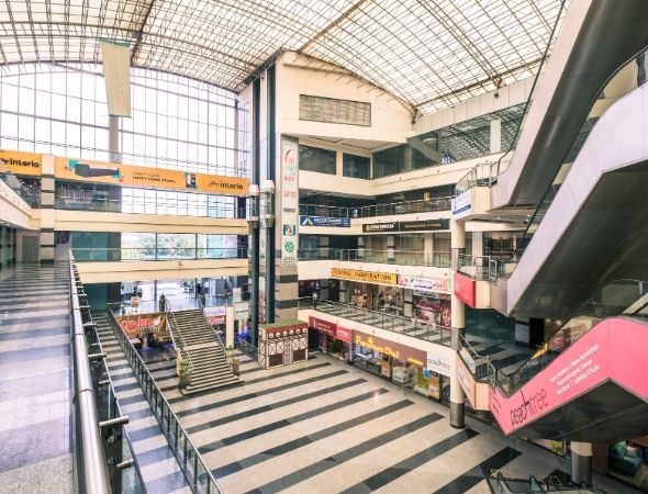 This is an indoor view of JMD Regent Arcade mall on MG Road Gurugram, showcasing a wide-angle perspective of its multi-story central atrium, featuring multiple levels of retail shopping corridors, moving escalators, and striped flooring under a large sloped glass skylight ceiling.