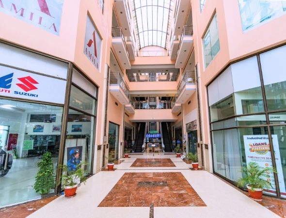 This is a wide interior perspective of JMD Regent Plaza on MG Road Gurugram, showing a long ground-floor retail alley lined with glass storefronts, multi-tier symmetry balconies, and a vaulted glass skylight ceiling providing natural day lighting.