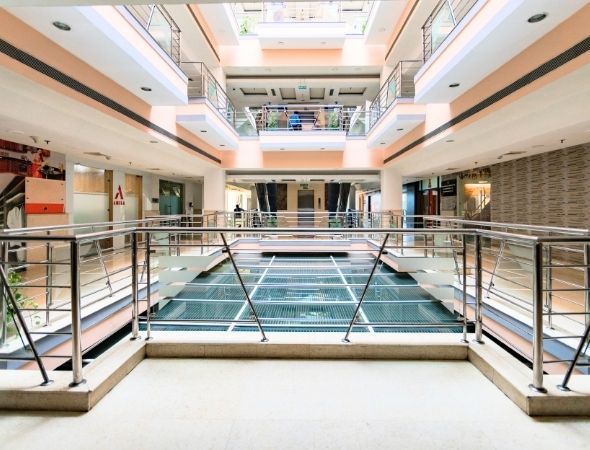 This is an interior architectural shot inside JMD Regent Plaza on MG Road Gurugram, showcasing a view from the upper floor corridors featuring modern steel and glass railings that look down into the open multi-story central atrium.