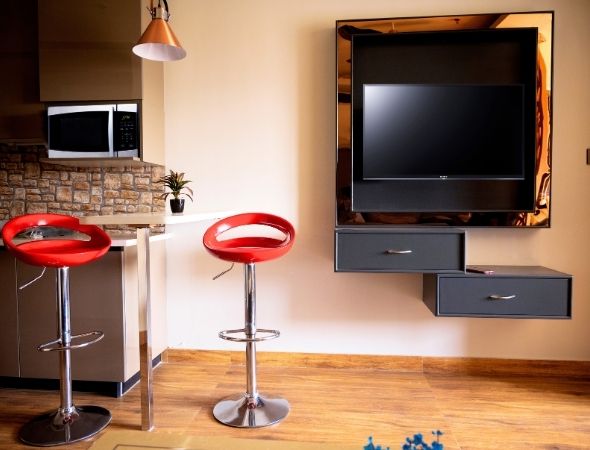 This is a JMD Suburbio 2 building in Sector 67 Gurugram showcasing a dual split view of a luxury studio apartment, and a modern kitchenette with red bar stools and a wall-mounted TV panel on the right.