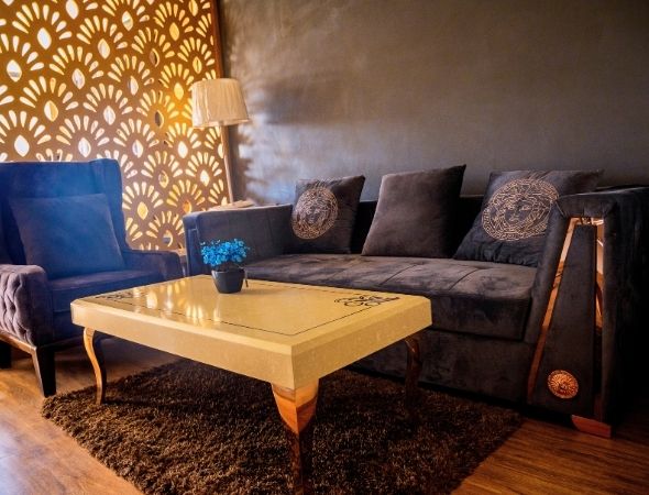 This is a JMD Suburbio 2 building in Sector 67 Gurugram showcasing a premium studio apartment living room setup, featuring a plush dark gray sofa set with patterned cushions, a marble-top coffee table over a shag rug, and a decorative wooden lattice partition screen.