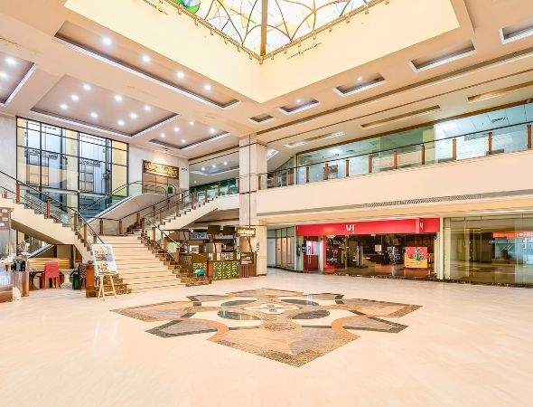 This is a wide-angle ground floor shot inside JMD Regent Square on MG Road Gurugram, displaying the spacious central lobby plaza with patterned marble flooring, multi-tier retail corridors, and brightly lit office entrance zones.
