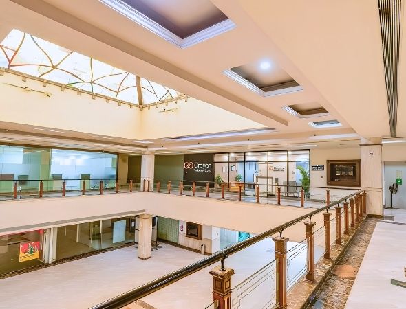 This is an indoor view of JMD Regent Square on MG Road Gurugram, showcasing the multi-level office corridors, a stylish false ceiling with square recessed panel lights, and a glass-railed balcony overlooking the central atrium