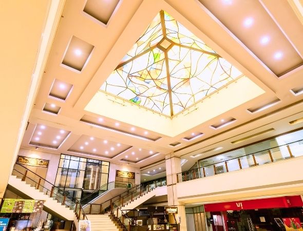 This is an interior architectural shot inside JMD Regent Square on MG Road Gurugram, featuring a grand pyramid-shaped glass skylight that floods the commercial atrium with natural daylight, framed by elegant ceiling lighting panels.